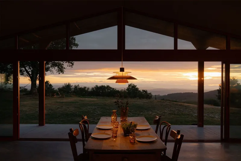 Dining table set beneath a tiered pendant light facing floor-to-ceiling glazing at sunset