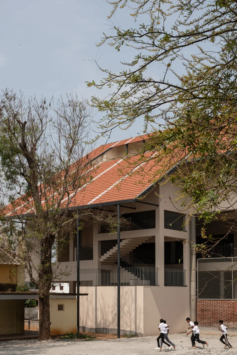 Corner view of the segmented terracotta roof volumes framed by trees with students running in the foreground