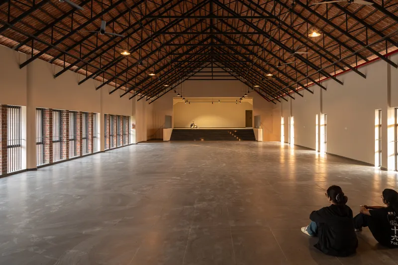 Multipurpose hall with exposed steel truss roof and concrete floor where two people sit