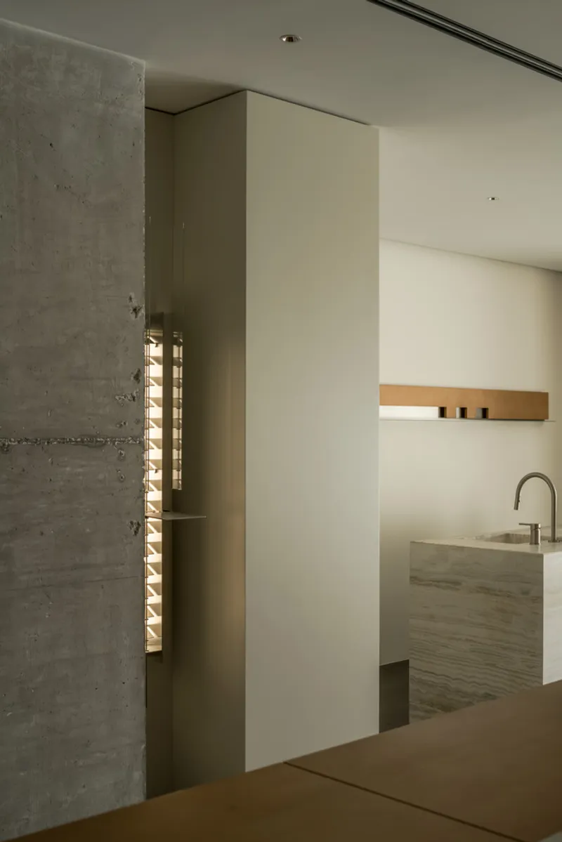 View through board-formed concrete wall to kitchenette with timber shelf and veined stone counter