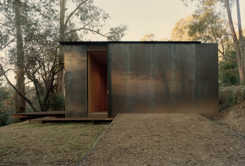 Weathered steel-clad entrance facade with narrow doorway at dusk among the trees
