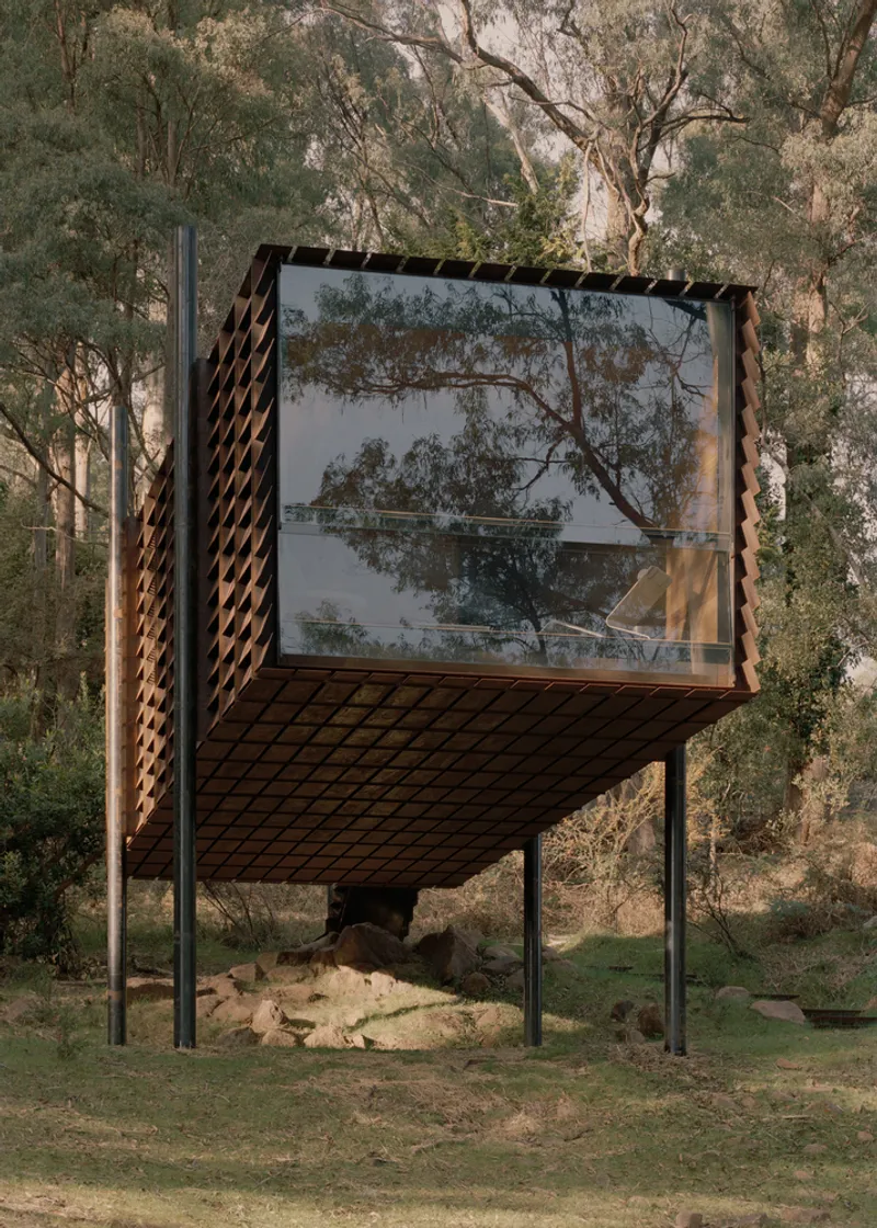 Glass-fronted box with diamond-patterned metal screens elevated above rocky ground in the forest
