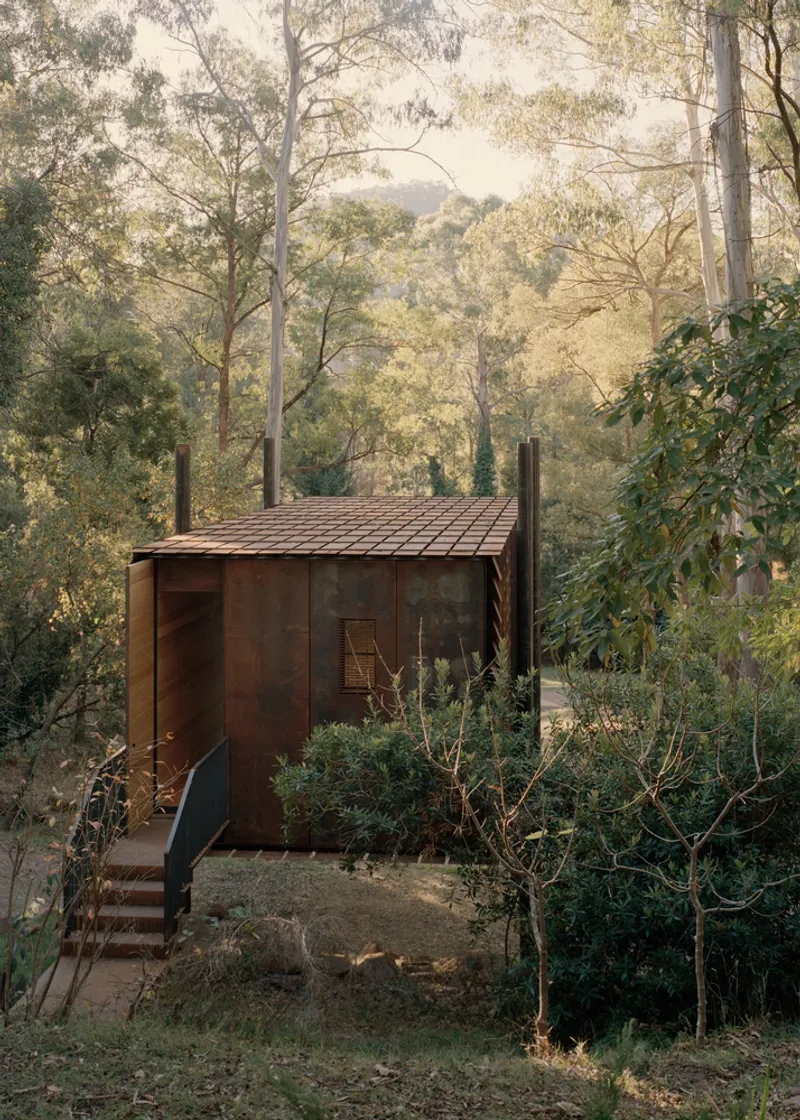 Weathered steel pavilion with tiled roof and external stair nestled among eucalyptus trees on a sloped site