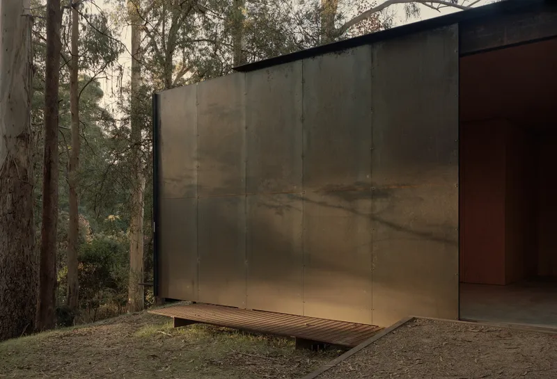 Weathered steel panel facade with timber deck ramp extending into a eucalyptus forest clearing
