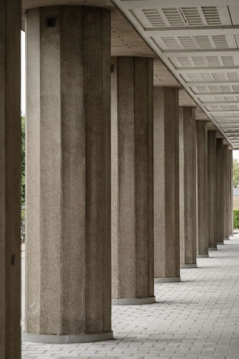 Colonnade of precast concrete columns with white perforated ceiling panels casting shadows on pavers