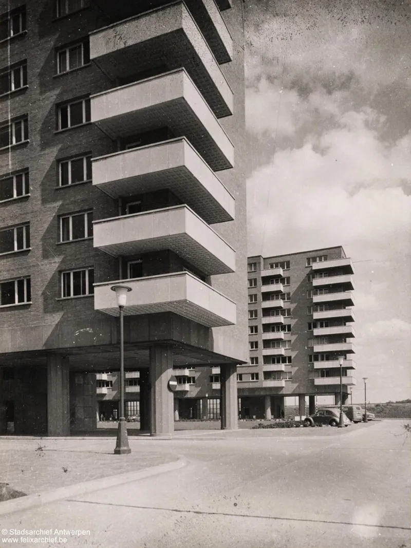 Street view of stacked balconies on pilotis with vintage cars and streetlamp in foreground