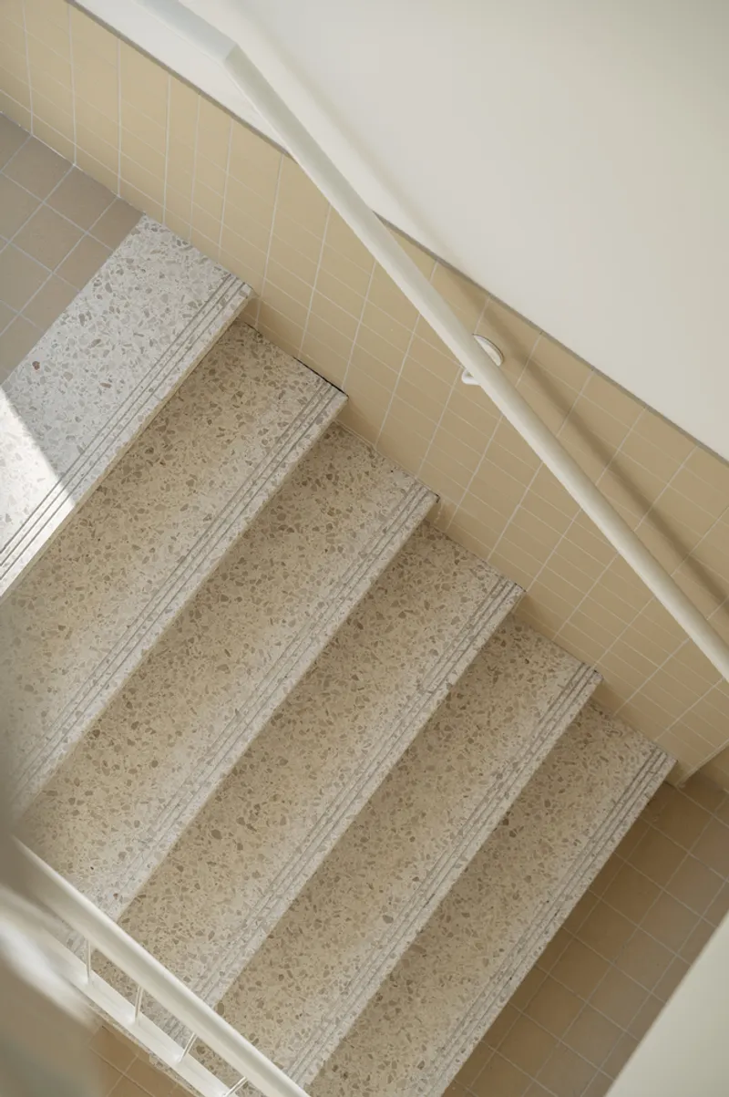 Top-down view of terrazzo stairs with grooved nosings between pale tiled walls and white handrail
