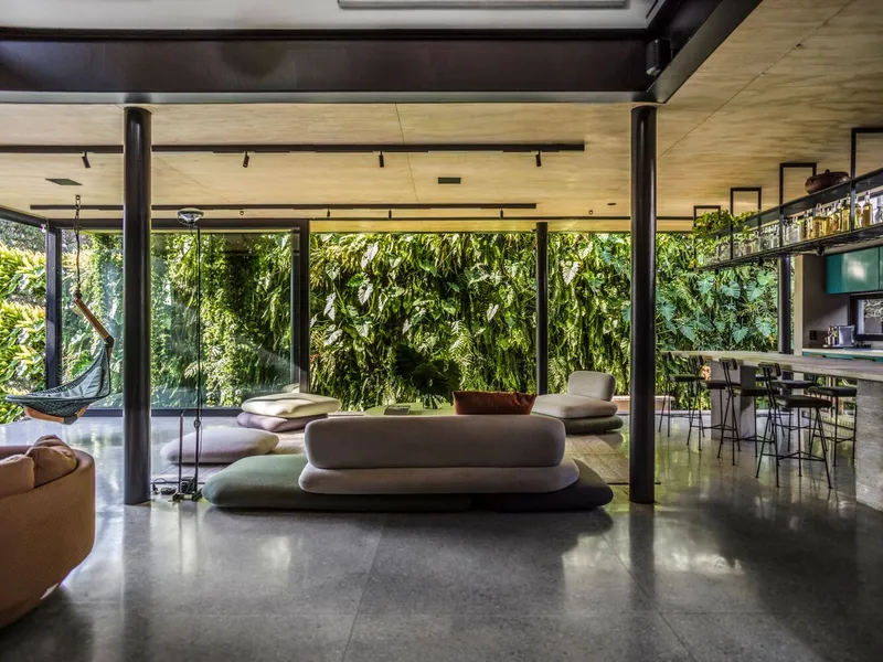 Open living area with steel columns and plywood ceiling facing a full-height green wall through glass