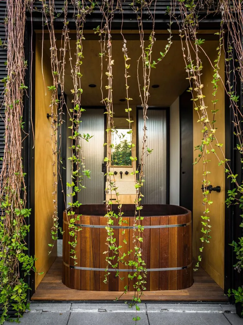 Timber soaking tub on a covered balcony with climbing vines descending from the slatted canopy above