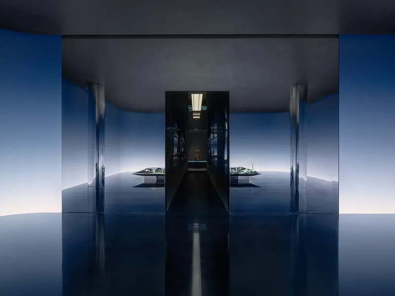 Symmetrical corridor with reflective blue floor and dark ceiling opening to a bright courtyard beyond