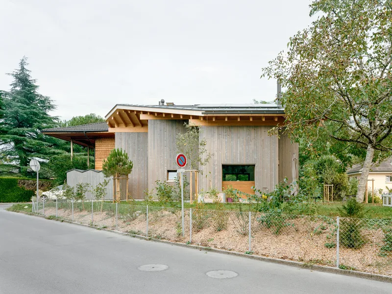 Street view of the gabled volume with chain-link fence and gravel lot in afternoon light