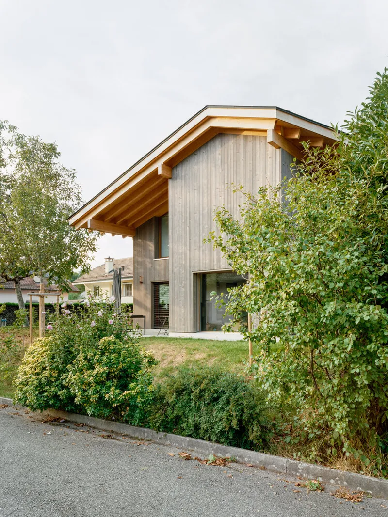 Gabled roofline with deep eaves framed by overgrown shrubs along the sloping street