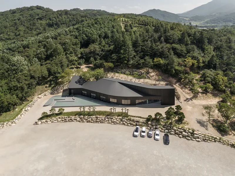 Aerial view of the black barn-form building set in a forested hillside clearing with parked cars