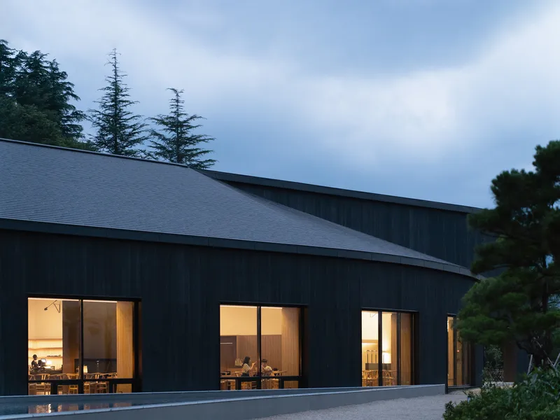 Dark timber facade with illuminated window openings revealing interior dining spaces at dusk