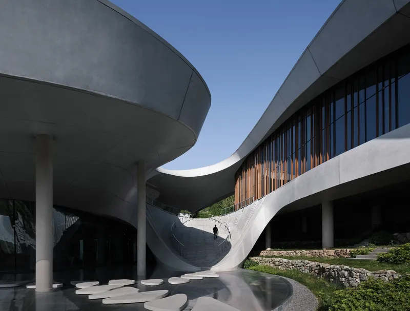 Sweeping white concrete staircase rising between vertical timber louvers with a lone figure ascending