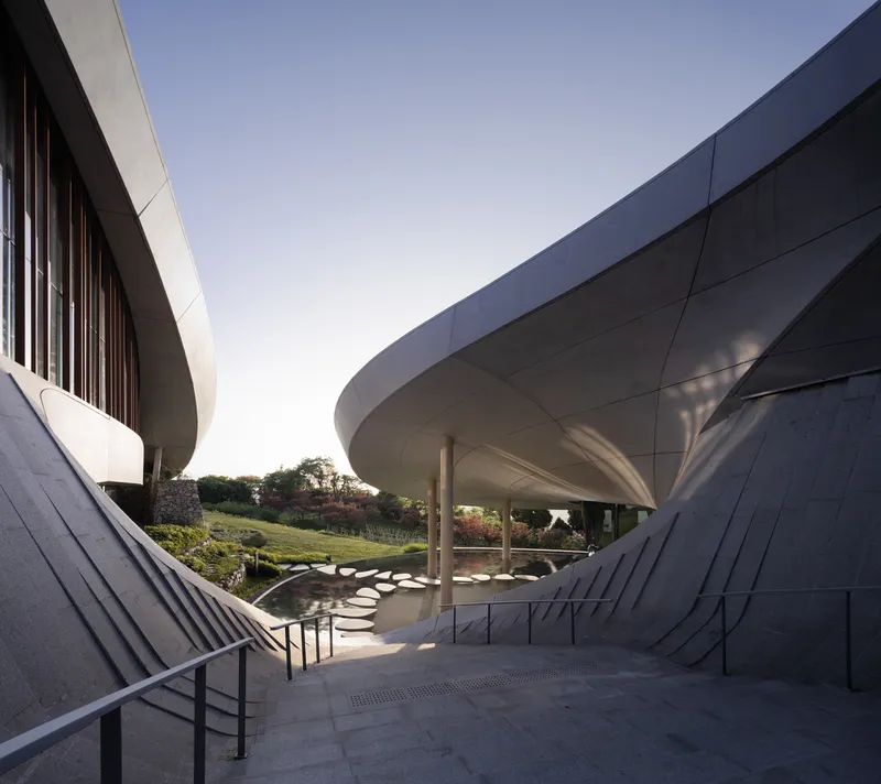 Entry staircase descending between folded panel walls beneath curving roof forms at dusk