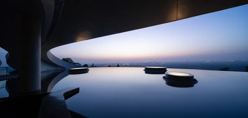 Infinity pool with circular platform beneath a curved concrete overhang at twilight