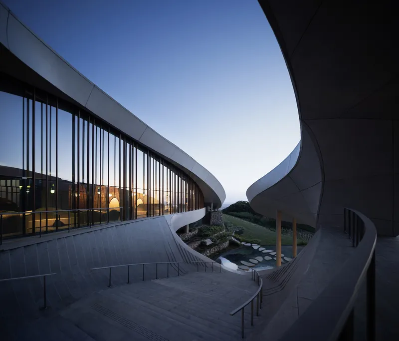 Sunken courtyard with planted bed between curved wings clad in vertical metal screens at dusk