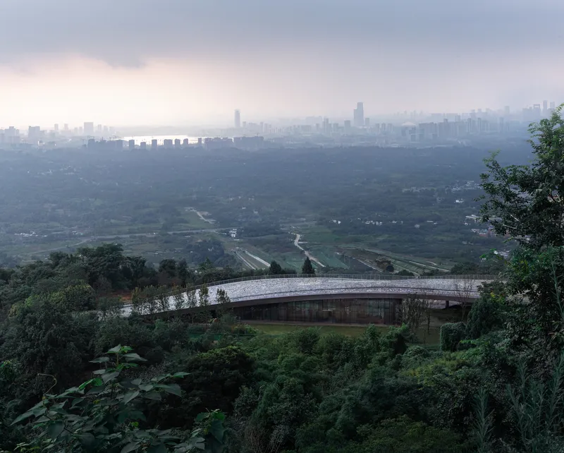 Low-slung roof structure nestled in dense green hillside vegetation with distant cityscape under hazy skies