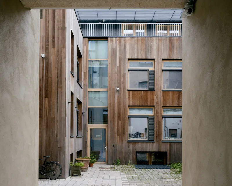 Narrow courtyard view of the vertical timber-clad facade with staggered windows and a rooftop balcony