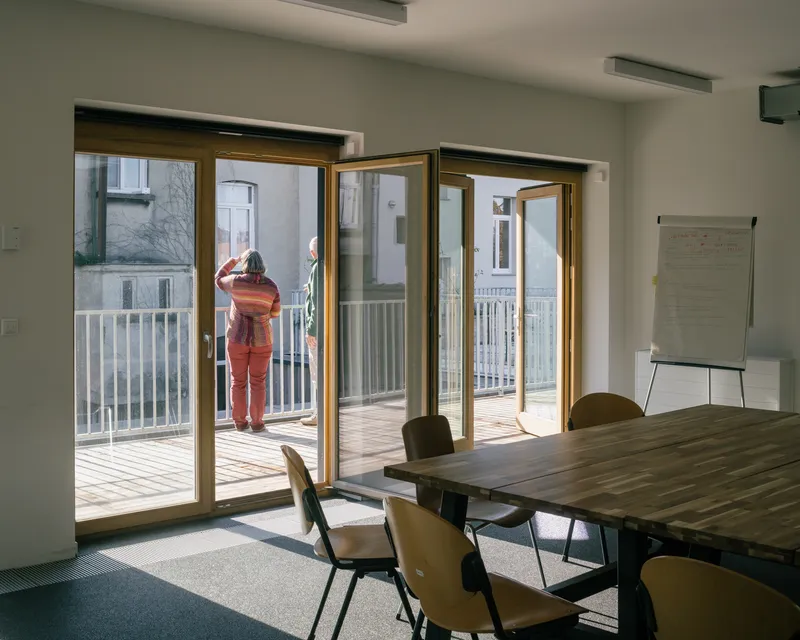 Interior view through timber-framed folding doors opening to a balcony where a person stands in sunlight