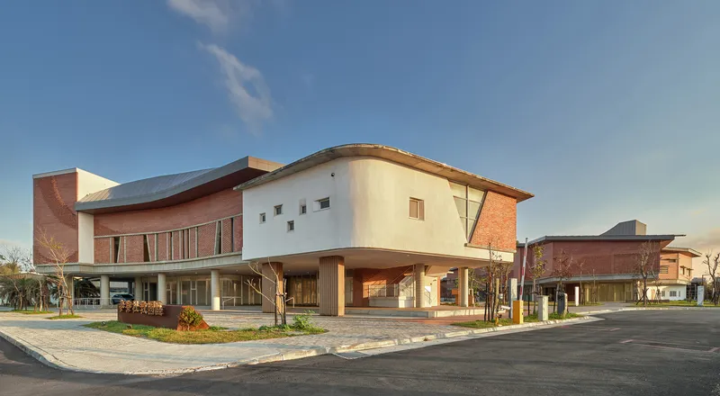 Street view of the complex showing curved and rectilinear volumes with red brick and stucco under evening light