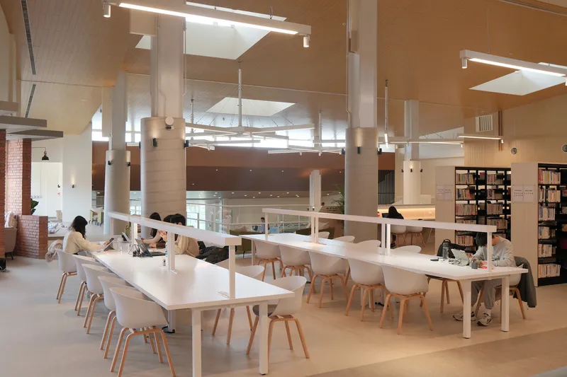 Double-height library interior with white study tables, wood chairs, and suspended linear lighting fixtures