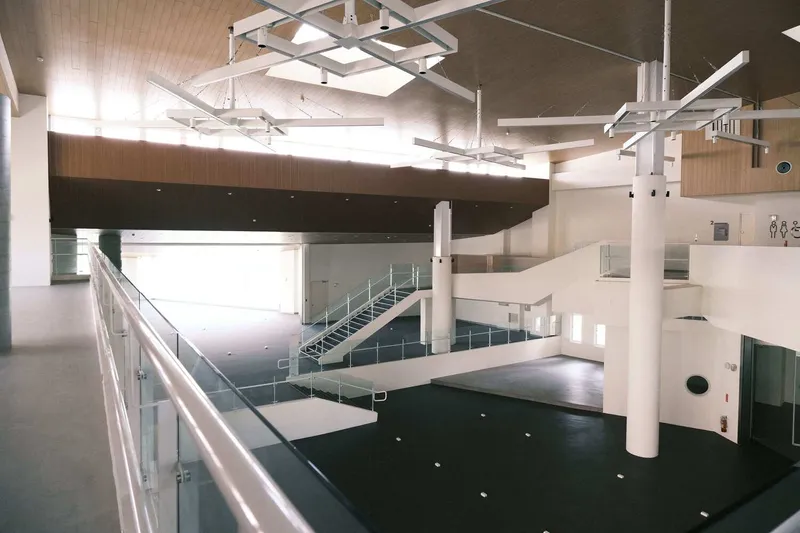 Glass-railed mezzanine overlooking black carpeted floor with white cylindrical columns and suspended linear light fixtures