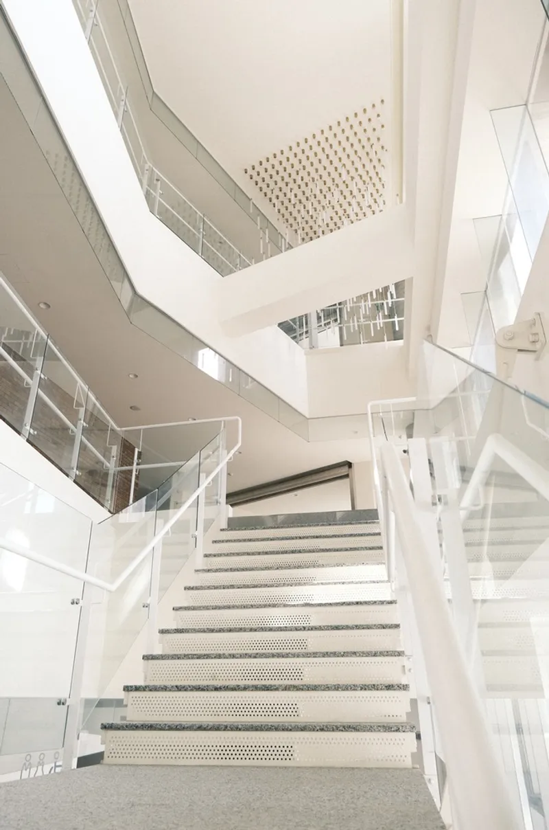 Perforated metal staircase rising through a multi-level atrium with glass balustrades and white walls