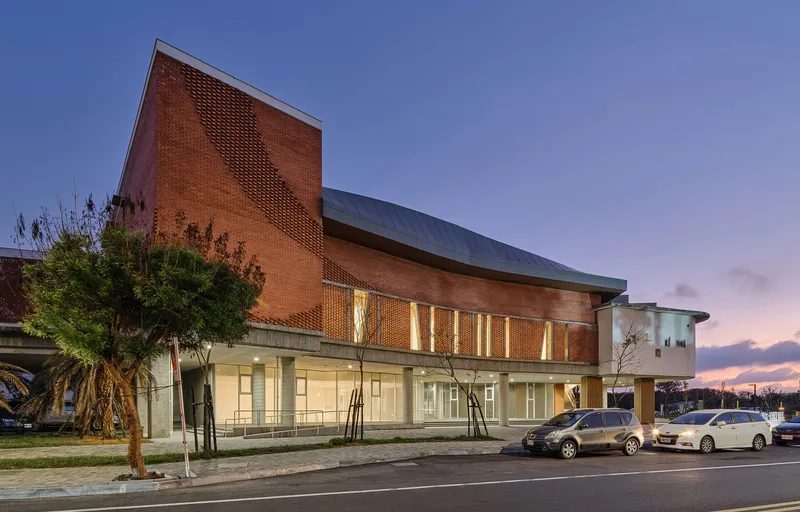 Brick facade with perforated screen tower illuminated at dusk beside a parking lot