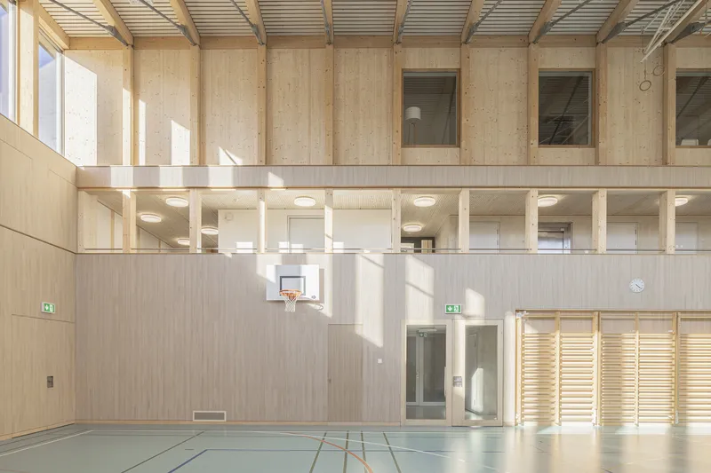 Interior gymnasium with exposed timber frame, mezzanine level, and clerestory windows casting sunlight