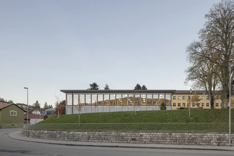 Glass-fronted pavilion atop a curved stone retaining wall with grassy slope in autumn light