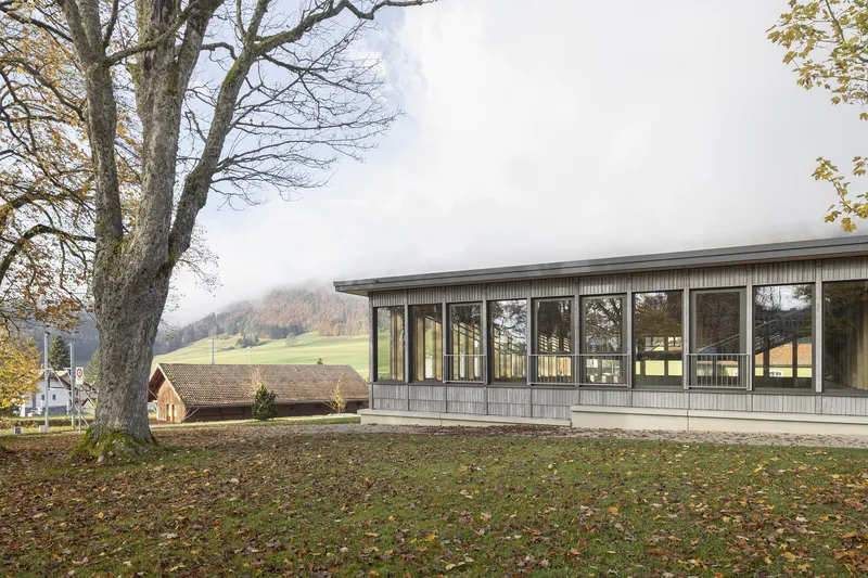 Glazed facade opening onto lawn with fallen leaves and misty hillside beyond