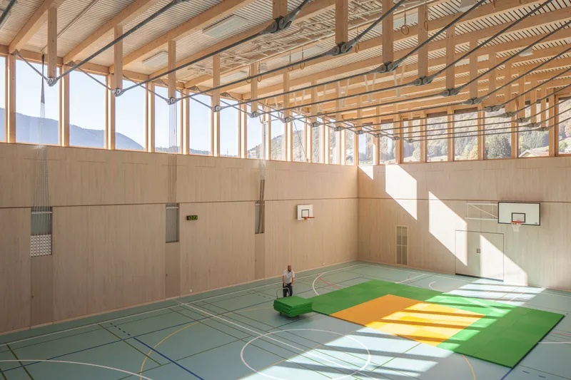 Gymnasium interior with a person seated on colored floor mats under strong afternoon sunlight through high windows