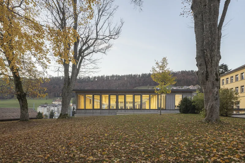 Single-story pavilion with floor-to-ceiling glazing illuminated at dusk among bare trees and fallen leaves