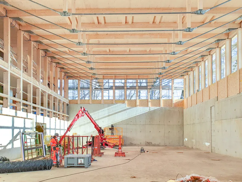 Interior construction view showing exposed timber beams and concrete walls with workers on scissor lifts
