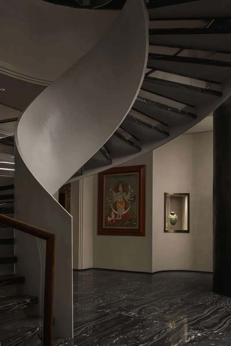 Helical staircase with white plastered wall curving upward past framed artwork in dim ambient lighting