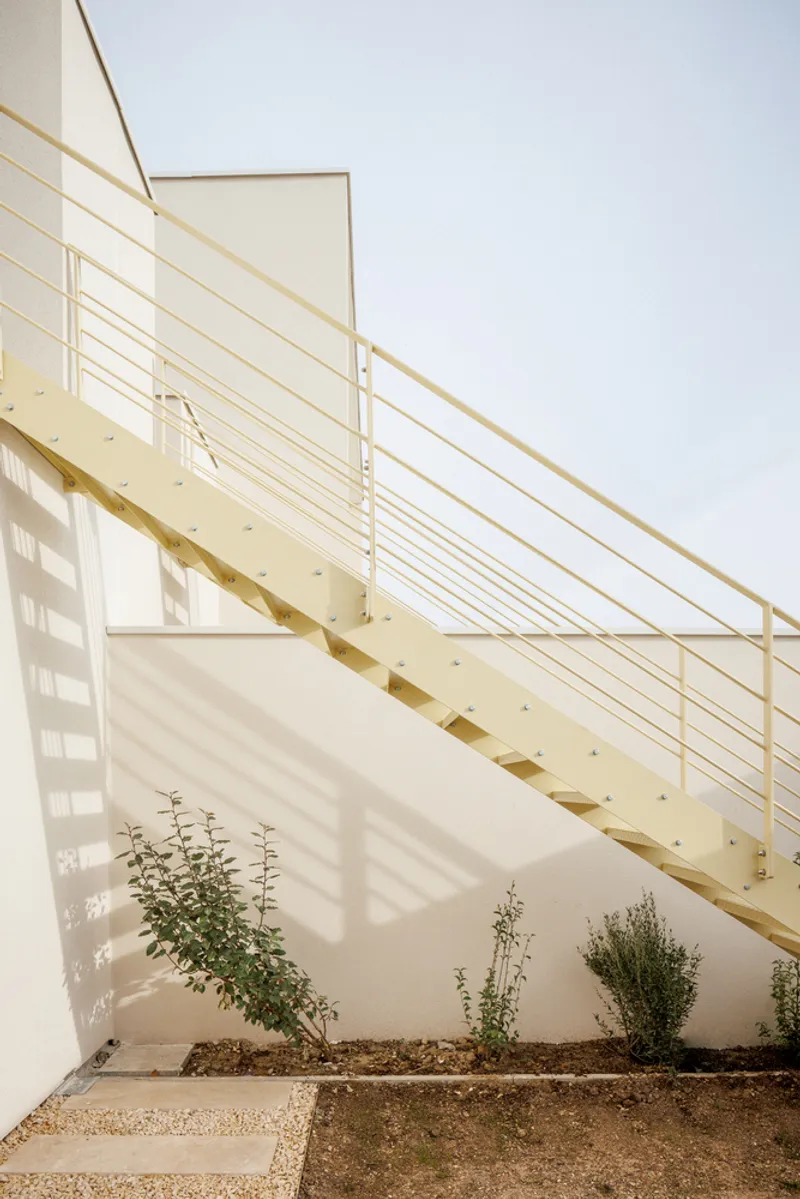 Exterior metal stair with horizontal rod railing casting shadows on a white wall with planted beds