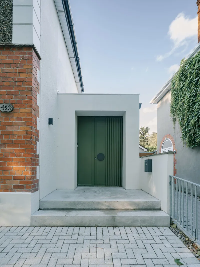 Street entry with dark green ribbed door and concrete steps between white rendered walls under blue sky