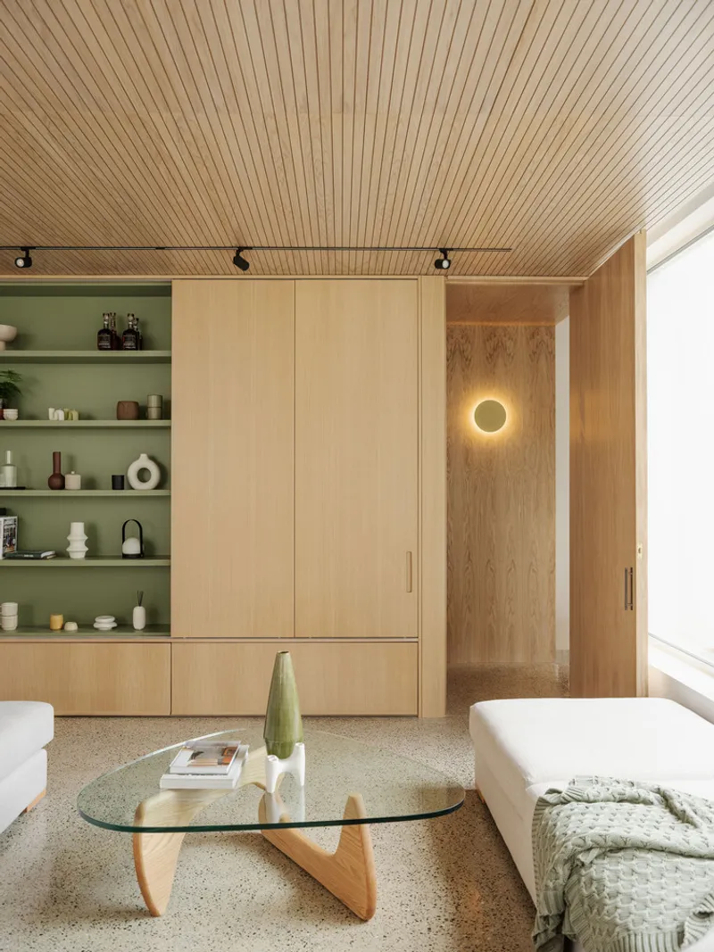 Oak storage wall with sage-green shelving niche and circular wall sconce beneath a slatted timber ceiling