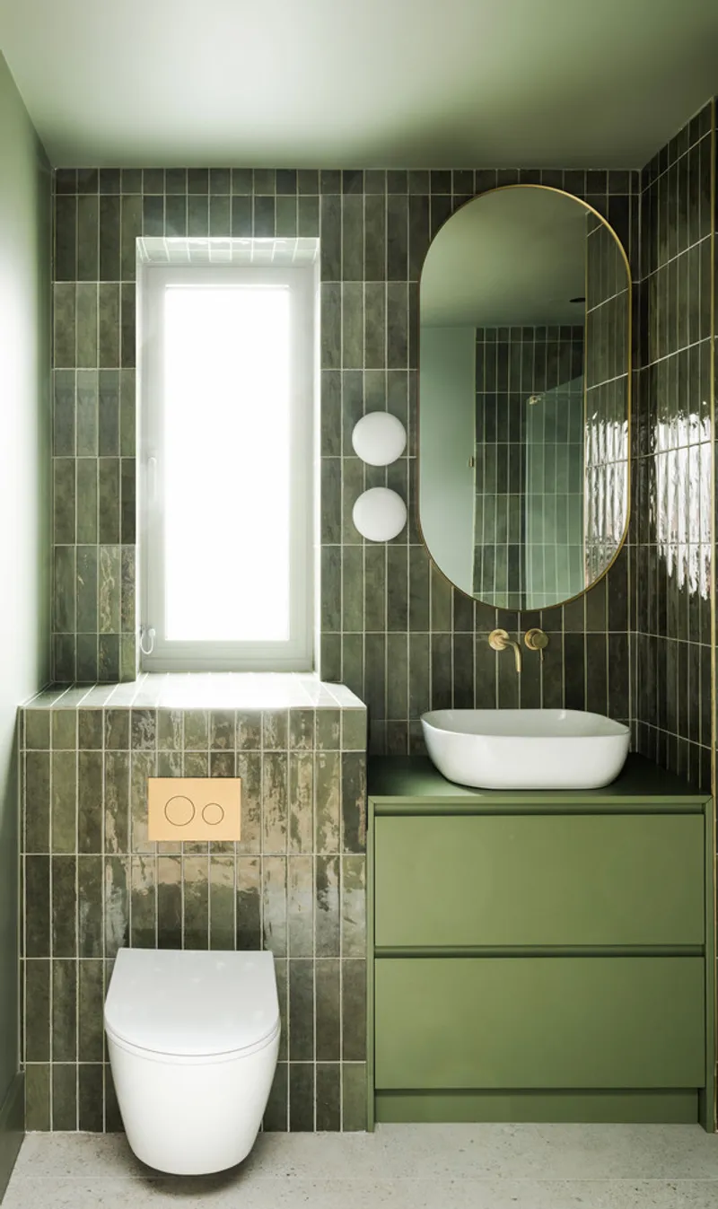 Bathroom with dark green vertical tile walls, round mirror, and wall-mounted toilet with gold flush plate