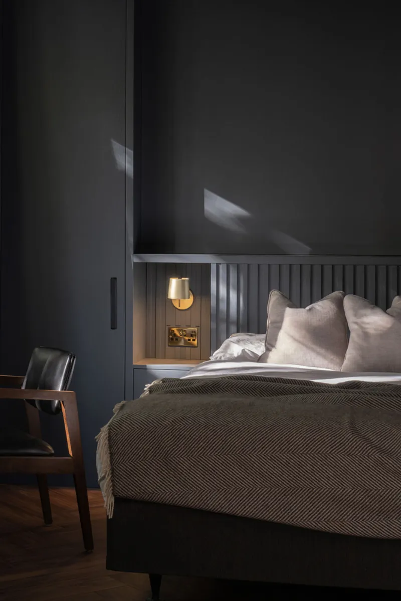 Bedroom with fluted headboard, wall-mounted brass sconce, and dark walls with soft window light