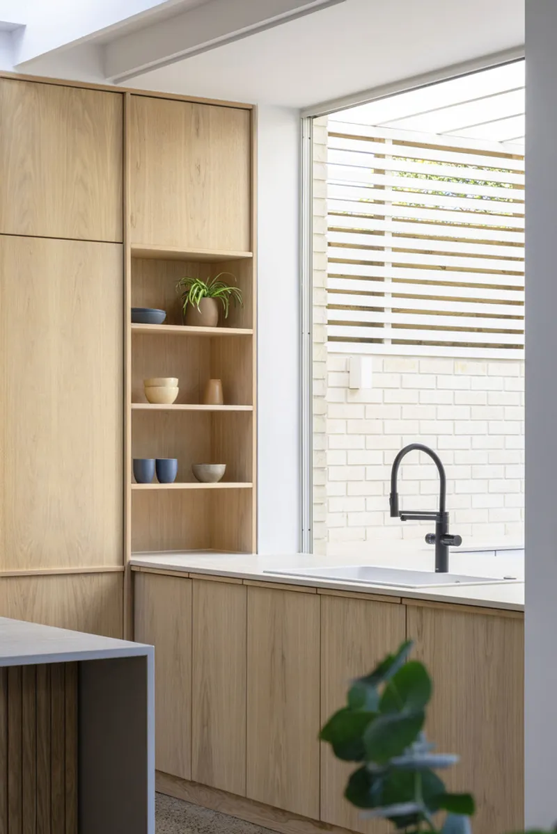 Kitchen cabinetry with open shelving displaying bowls and plant, white brick courtyard visible through louvred window
