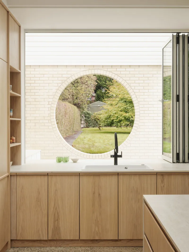Kitchen with light timber cabinetry and circular window framing a view of the garden beyond