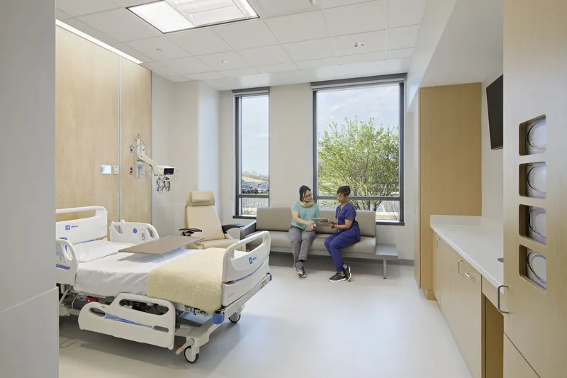 Patient room with hospital bed, seating area, and windows overlooking spring foliage
