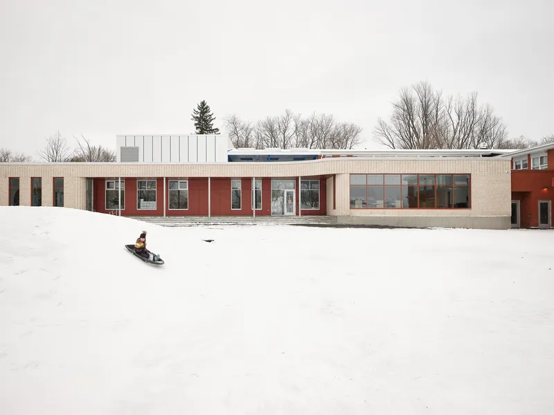Long low facade with alternating red and cream brick panels as a child sleds across the snowy yard