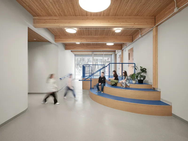 Corridor with exposed timber beams and curved wooden seating platform beside a blue metal staircase