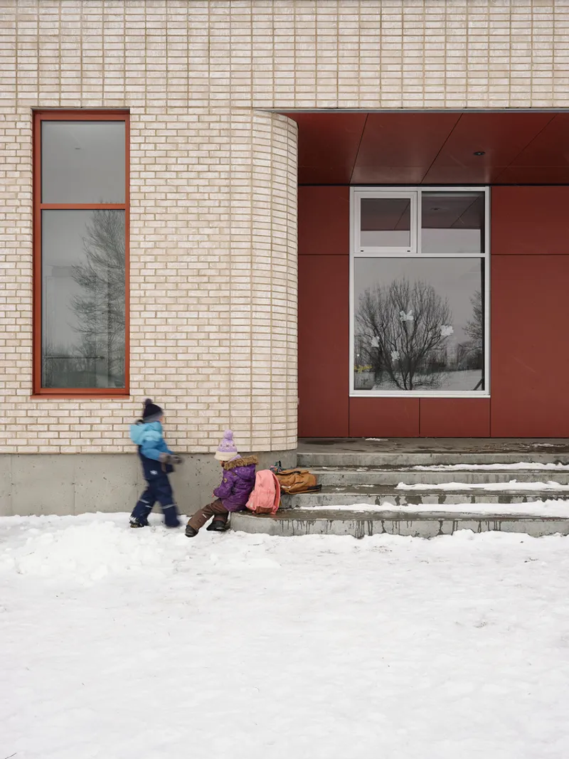 Curved brick wall meeting a maroon panel facade with children playing on snowy steps outside