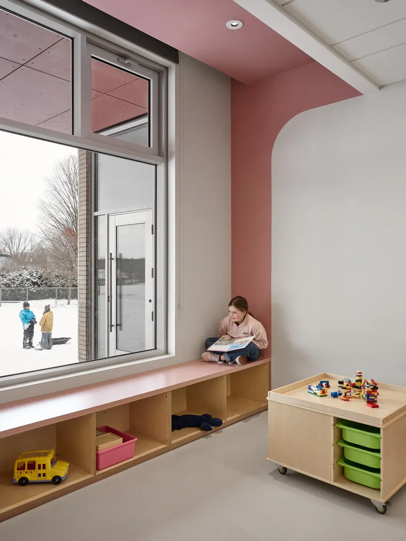 Reading nook with built-in timber bench and pink ceiling panel beside a window overlooking winter playground