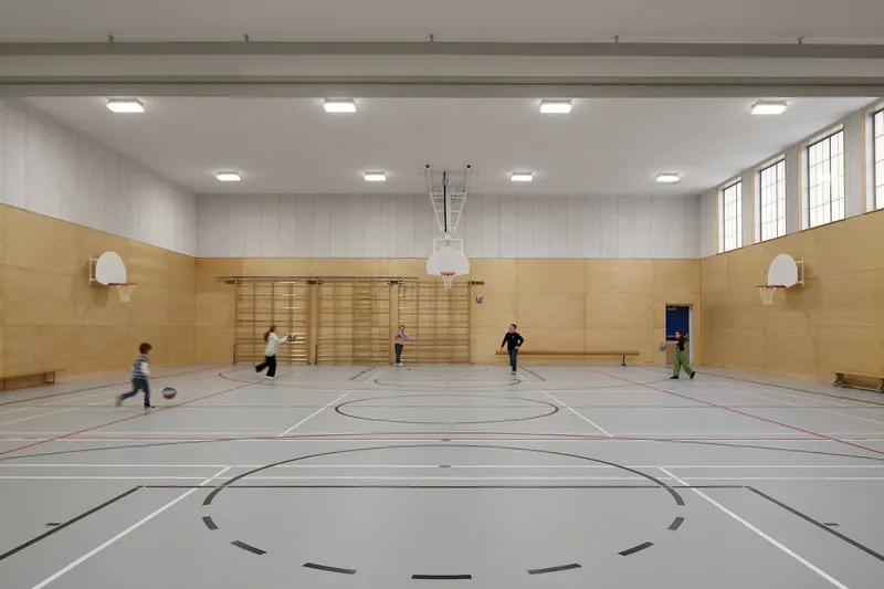Indoor sports hall with timber wainscoting and wall-mounted basketball hoop where children play ball games